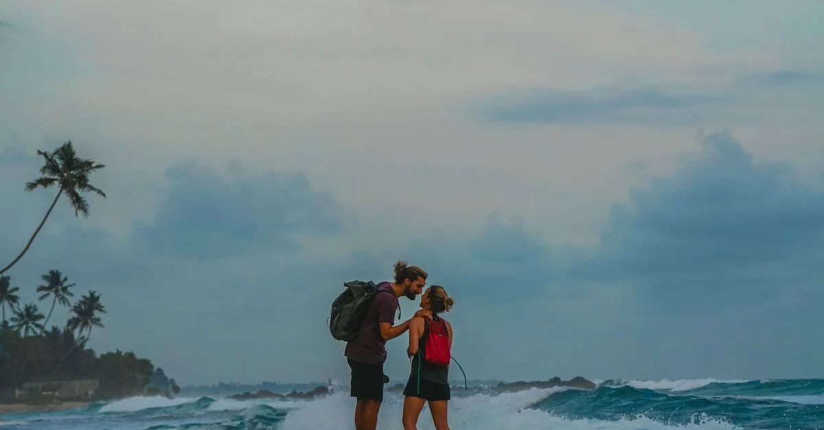 Un couple avec des sacs à dos s'embrassant sur une plage tropicale alors que les vagues éclatent, transmettant amour et aventure.