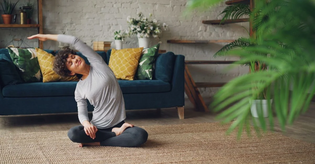 Femme exécutant une pose de yoga sur un tapis dans un salon confortable avec des plantes.