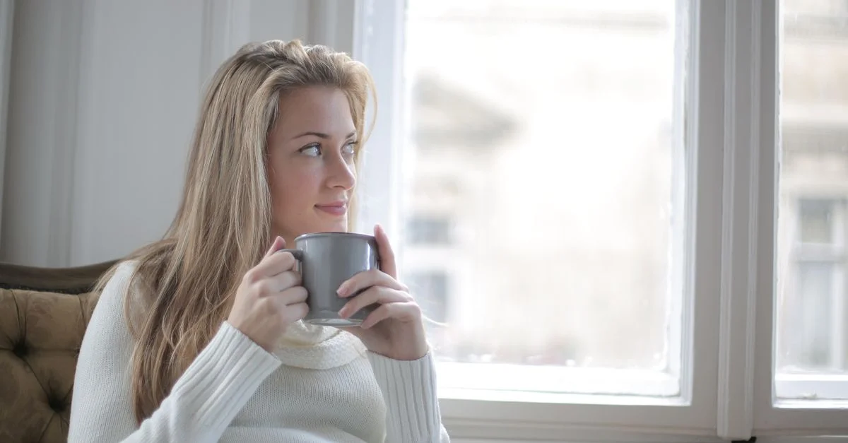 Une femme dans un pull confortable dégustant un café près d'une grande fenêtre ensoleillée.